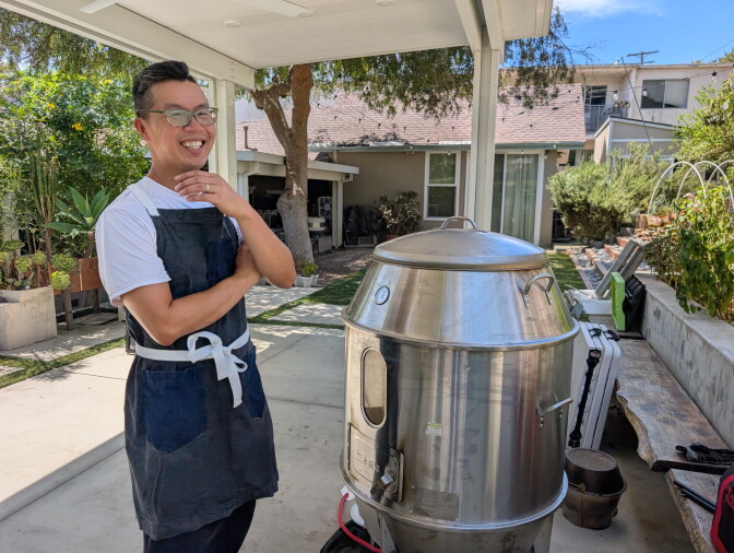 An Asian man in glasses and an apron smiling next to a spaceship-like outdoor oven.