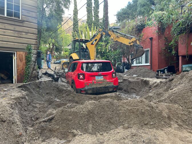 A red car next to two homes is engulfed in mud from a storm.