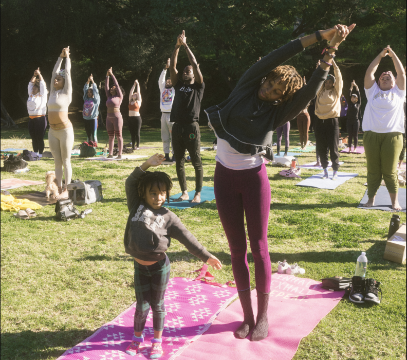 An adult and child, both with dark tone skin, engage in a yoga pose in a park with other participants behind them. They're on pink mats.