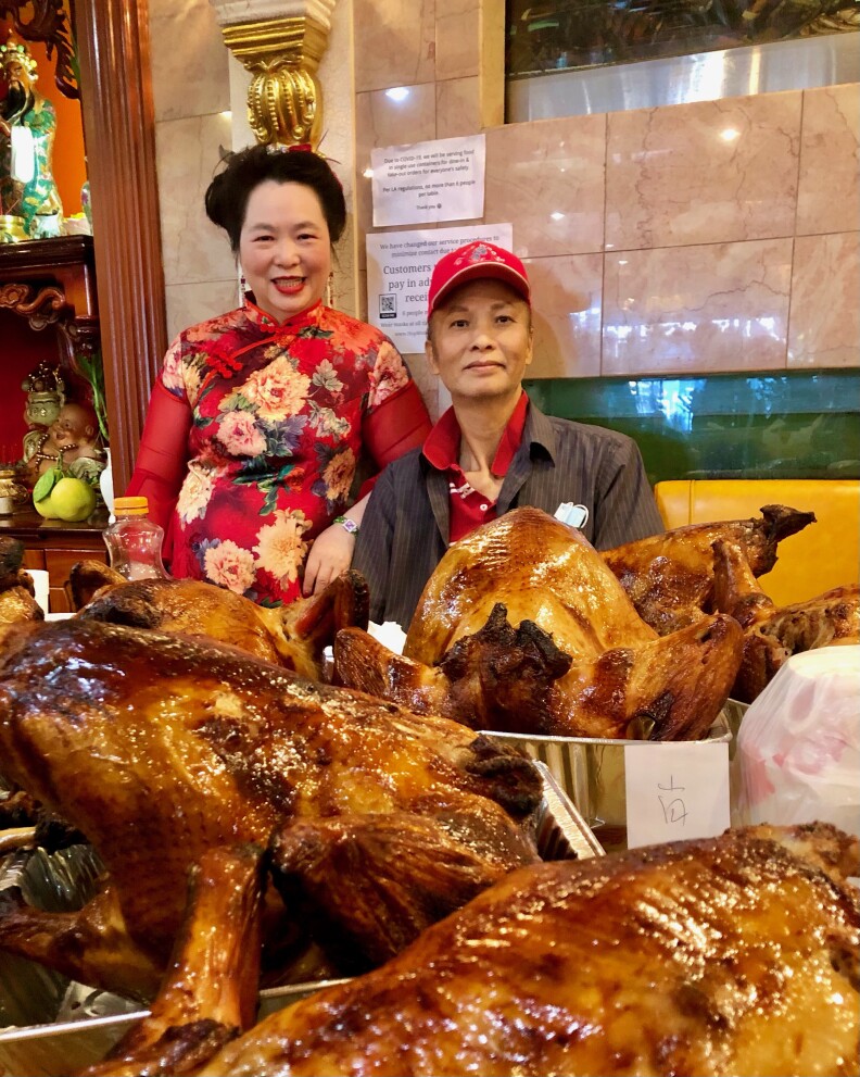 An older man and woman sit or stand behind a table laden with glazed roasted turkeys