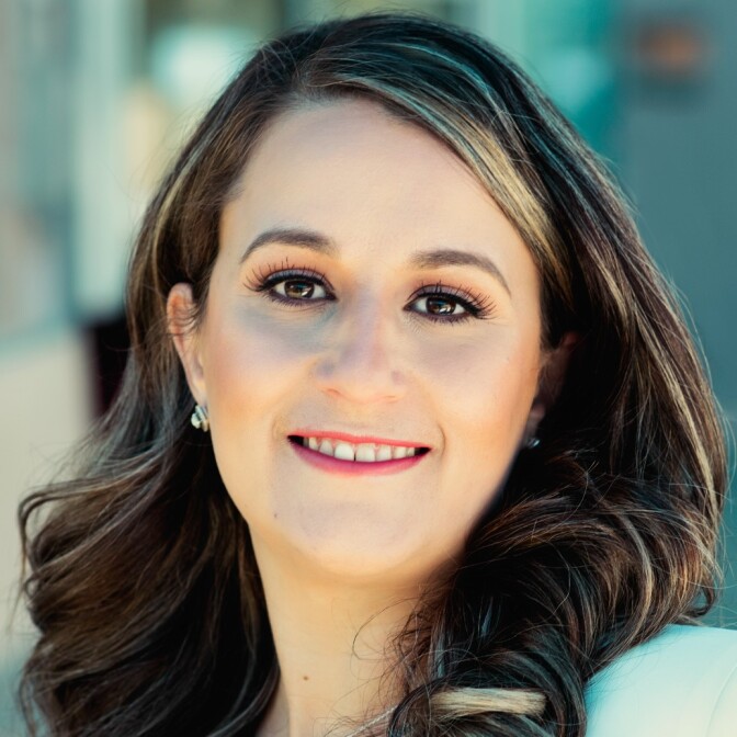 Imelda Padilla, a light-skinned Latina, smiles at the camera. She has brown eyes, and dark hair with a few blond streaks in it. 