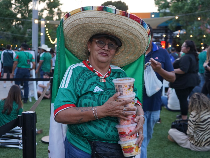 A medium-dark skin toned woman poses in a large sombrero and green Mexico jersey. She wears glasses and carries a stack of plastic containers full of food. A crowd of people are behind her.