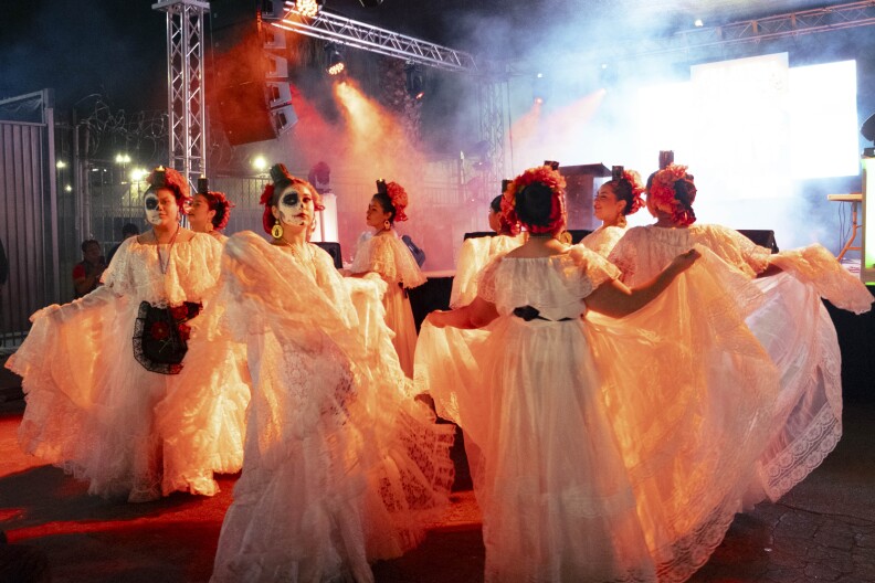 A group of multiple young women dance in front of the stage. They are wearing traditional white folklórico dress with intracate lace patterns.