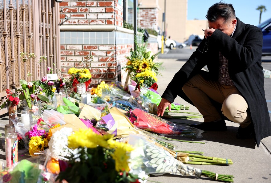 An Asian man in a dark suit crouches on a sidewalk stacked with flower bouquets. His hand is raised to his eyes in emotion.