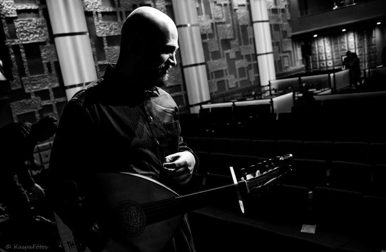 A black and white photo showing a bald man holding a stringed instrument looking towards towards the ground. There are tables and chairs in the background.