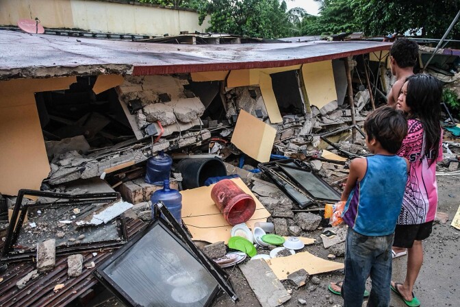 A man and two children stand in front of the remains of a collapsed building