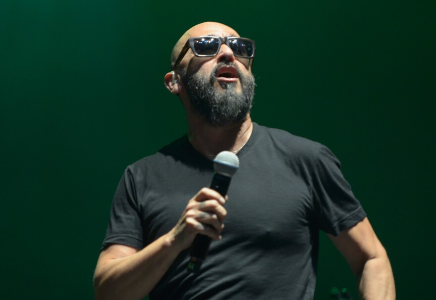 A man with light brown skin wears a black t-shirt and sunglasses sings into a microphone.