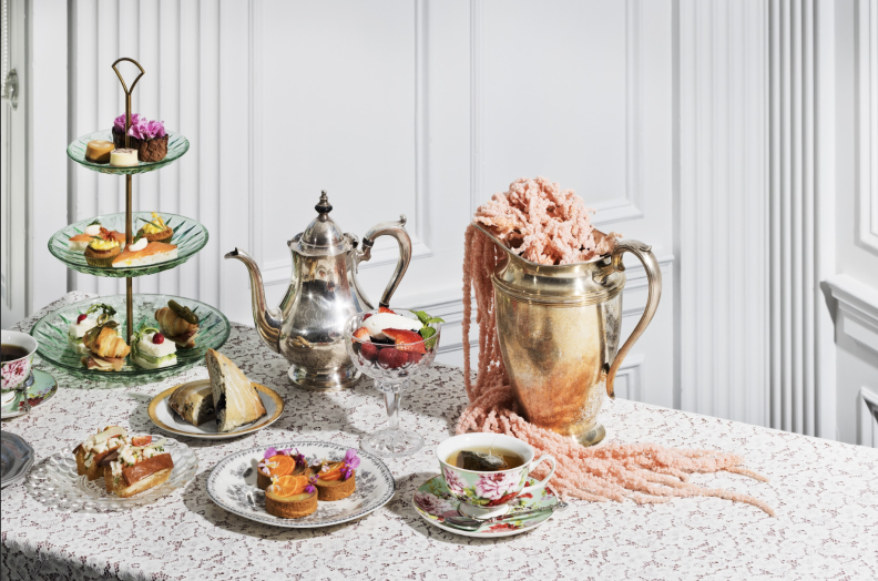 A table is set for tea, with a towering glass-and-silver tray for finger foods, a silver tea pot, a silver vase strewn with flowered vines, and an assortment of dainty dishes and tea cups. It all sits on a white tablecloth that is adorned with tiny pink flower.