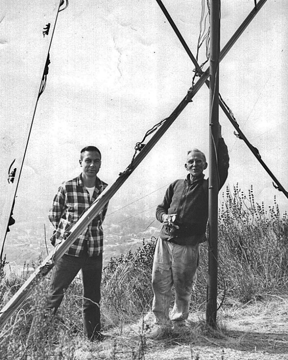 Two man standing next to a wood structure, with lightbulbs on some of the beams.