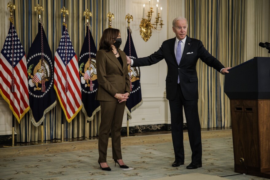 President Biden, on a dark suit and light purple tie, has one hand on a lectern and the other on the left shoulder of Vice President Kamala Harris, who where an oliove green pantsuit and a black mask. Behind them are U.S. flags and flags with the presidential seal.