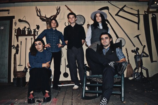 The five members of the band Darden pose together in a room with a wooden floor and instruments and metal tools on a wall behind them.