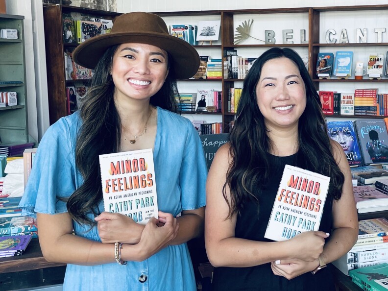 Two Asian American women -- one woman on left is wearing a wide-rimmed hat and a denim outfit, woman on right is in a sleeveless black top -- each hold copies of the book "Minor Feelings" by Cathy Park Hong.