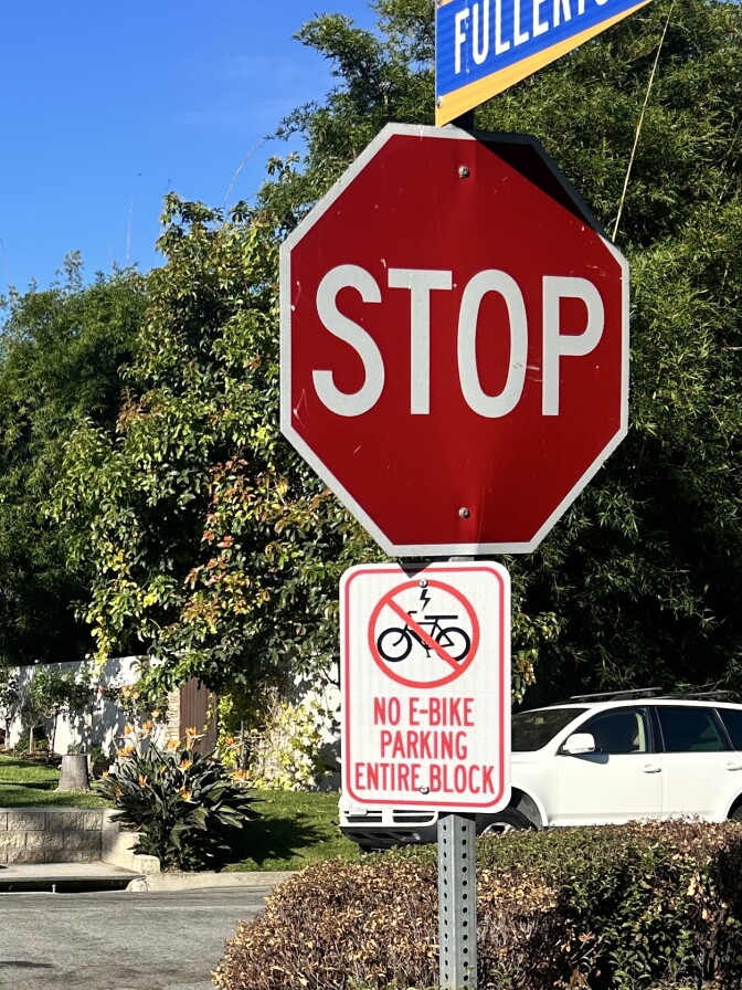 A stop sign stands above a placard reading 'No e-bike parking entire block,' with a Fullerton street sign partially visible overhead.