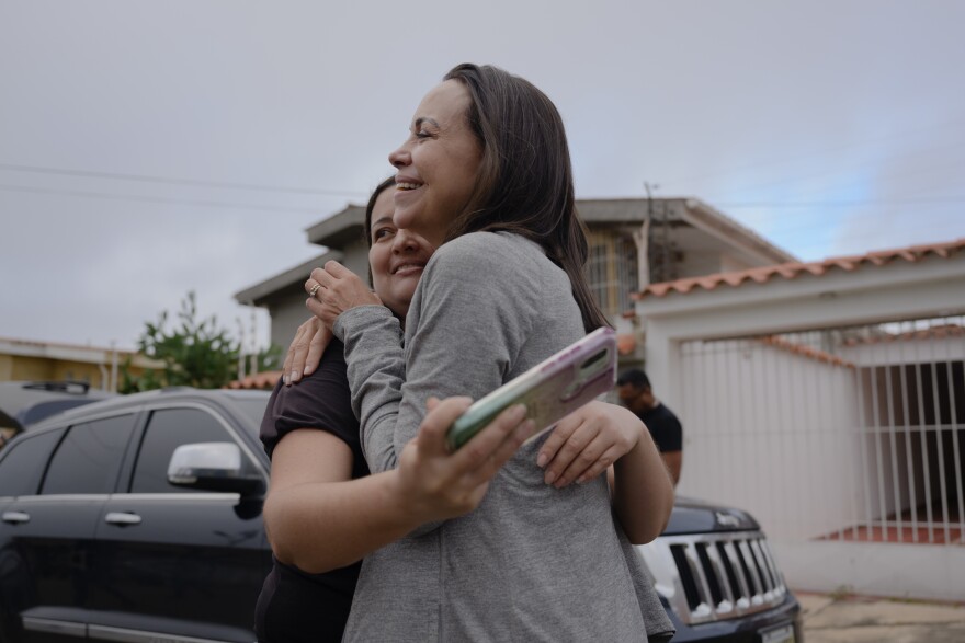 A woman wearing a black T-shirt embrace another woman wearing a grey sweater. They are standing on the street in front a parked black SUV and a house with a white, metal gate. The woman wearing the black T-shirt holds a cellphone in her right hand