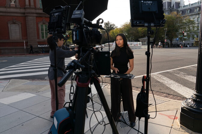 A woman wearing all black stands on a street corner in front of a television camera.