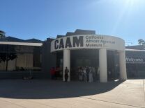People gather in the shade under the sign for CAAM, the California African American Museum.