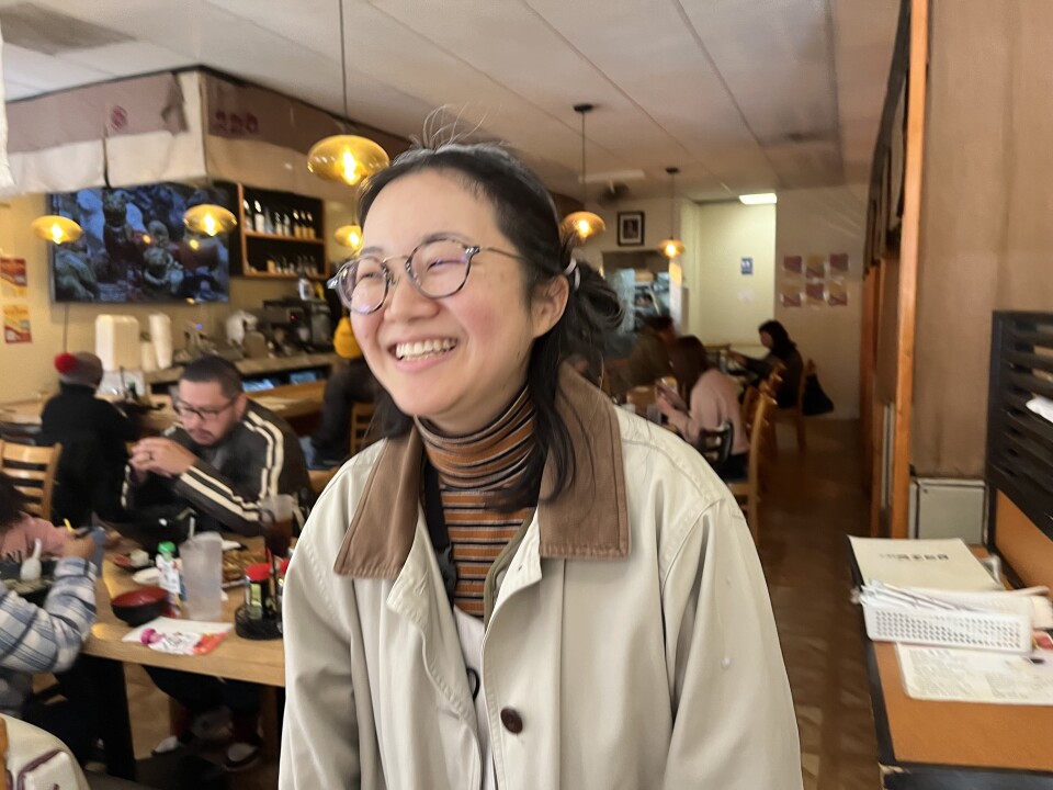 A Japanese woman in a beige barn coat beams while standing in a busy Japanese restaurant.