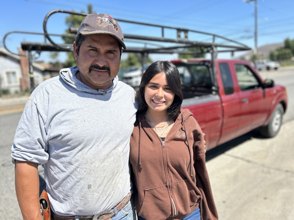 A middle-aged man with light brown skin and a black msutache and grey sweatshirt stands with his 15-yearold daughter who has long black hair, wears a brown sweatshirt. They both smile. It's sunny and a red truck is blurred behind them. 