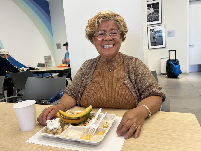 A Latina woman wearing a brown sweater over a shirt in a lighter shade of brown. She's sitting at a table, with a lunch tray and two slices of cantaloupe placed on top of the tray. A white paper cup is sitting next to the tray.