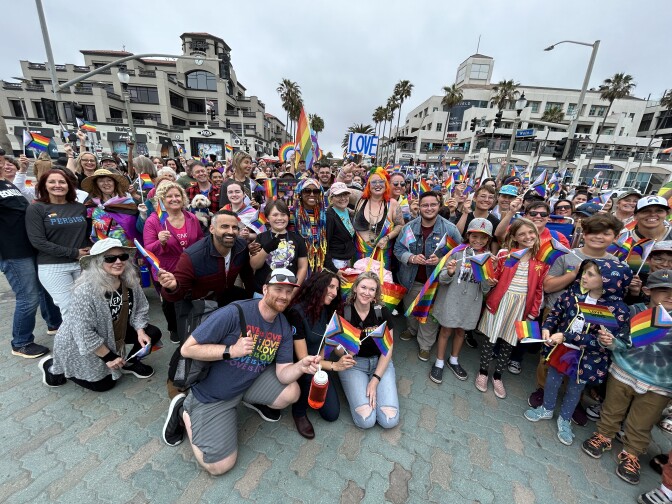 A crowd of people, roughly 50 to 100, huddle for a group photo. People are different ages, young and old, with varying skin tones. Most people have pride flags that they're waving in the air.