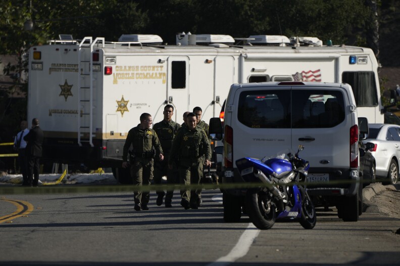 A group of law enforcement officers in green tactical gear walks along a road that has been blocked off by yellow police tape. On the side of the road is a white van and a blue motorcycle, and behind them is an RV-sized vehicle labeled "Orange County Mobile Command" and with a gold sheriff's star painted on the sides.