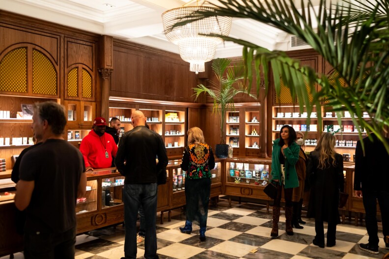 A room with wood paneling and shelving full of various cannabis supplies. Various people stand around the counter that wraps around the room with a black and white checkered floor.