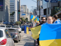 Several people stand on the side of a busy urban street holding Ukrainian flags.