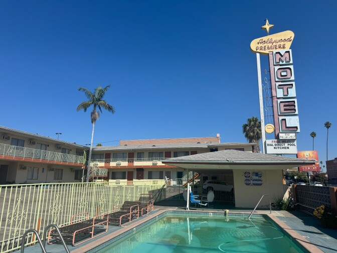 Wide shot of the Hollywood Premiere Motel showing a two-story building with mint green railings, a turquoise rectangular swimming pool in the foreground, and the iconic tall white and pink "MOTEL" blade sign with starburst design against a clear blue sky.
