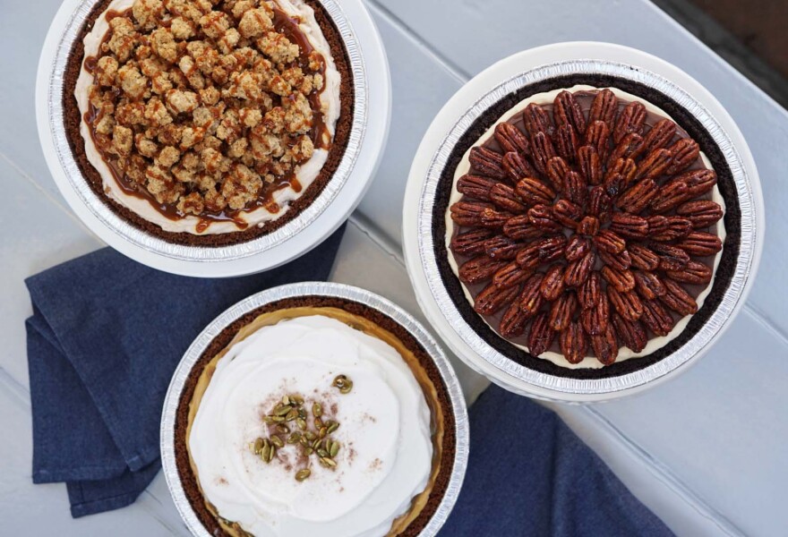 An aerial view of three ice cream pies in white tins on a white picnic table, and with two blue napkins folded underneath them.