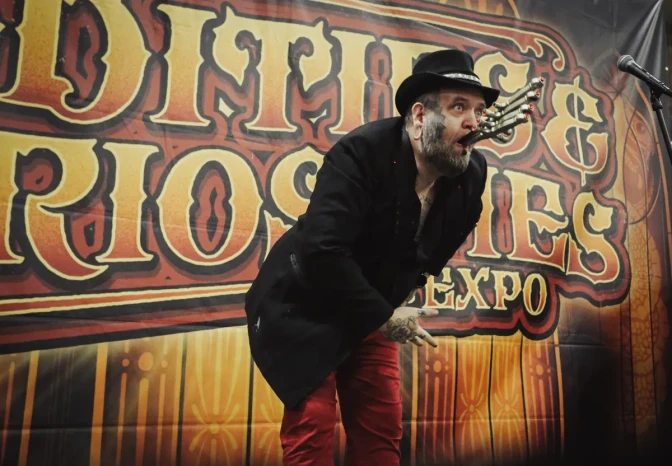 A light-skinned man with a beard and black hat holds a strange metal contraption in his mouth. 