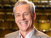 Michael Ritchie, in a portrait, smiles in a light-colored suit with his arms crossed.
