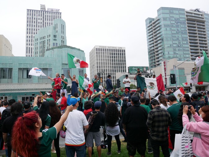 A group of people are standing outside watching another group of people holding the Mexican and Korean flags.