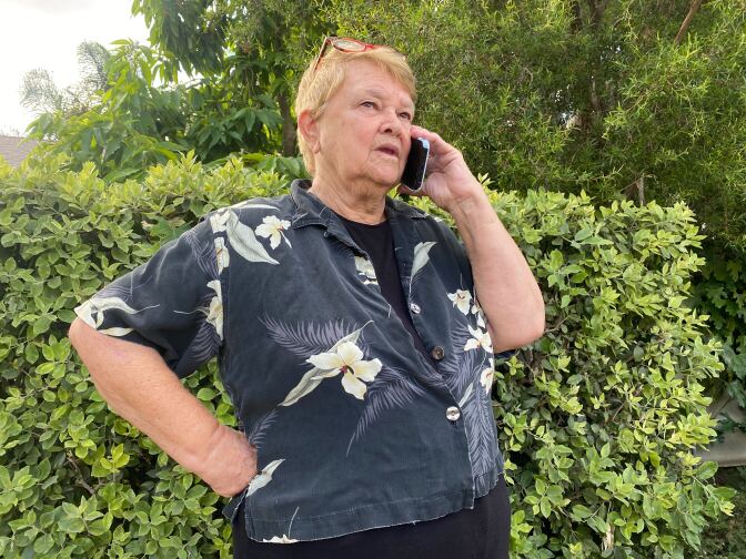 Supervisor Sheila Kuehl, a white woman with short blond hair wearing a dark blue shirt with white flowers and leaves, talks into a cell phone she holds with her left hand, with her right hand on her hip. 