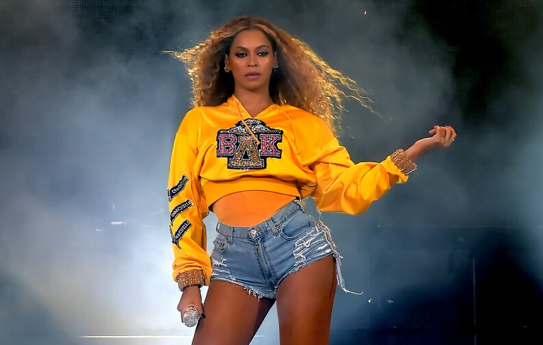A woman with dark-tone skin and flowing hair wears a yellow satin-y cutoff jersey with a glittery sorority logo on it, jean shorts, and holding a microphone in front of fog and dramatic lighting.