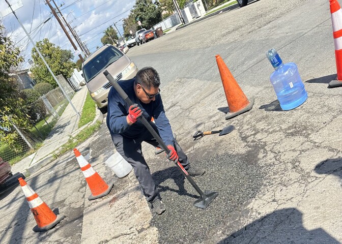 A man with a medium dark skin tone stands on a street outside leaning down to stomp the ground with a tool. The tool is flat at the bottom to compact the aphalt that was freshly laid down over a damaged road area.