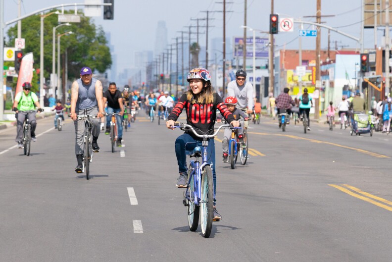 People ride down a street on bicycles with the DTLA skyline behind them