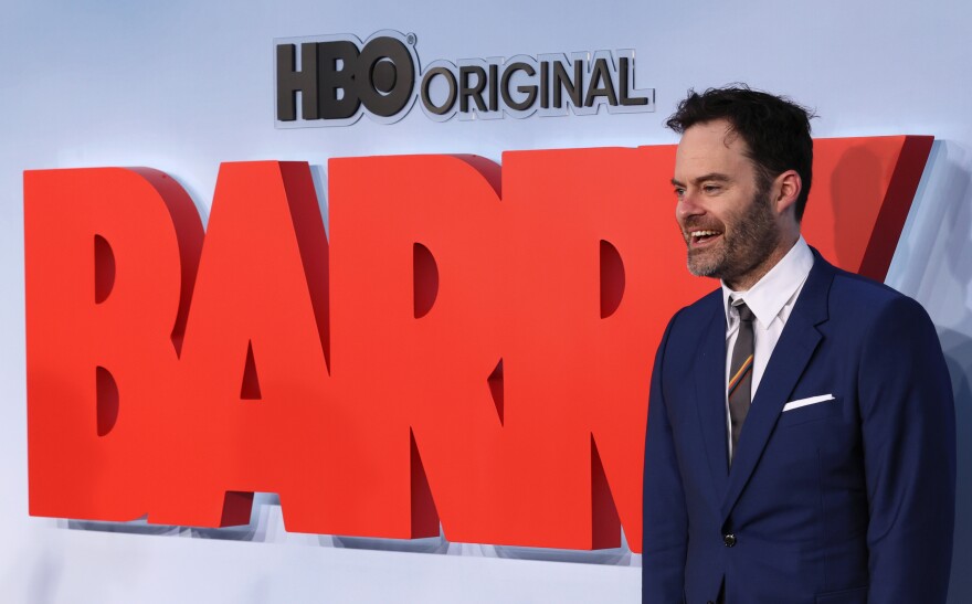 This premiere-arrival shot shows a man with light skin tone in a blue suit standing next to a sign that reads "BARRY" in big, reddish-orange block letters. Standing on the right of this image, he is looking and smiling to the left, presumably at a volley of onlookers and press.