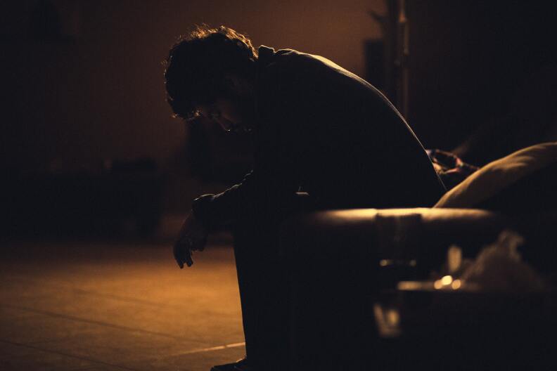 A man is silhouetted as he sits on a bench, looking down and dejected.