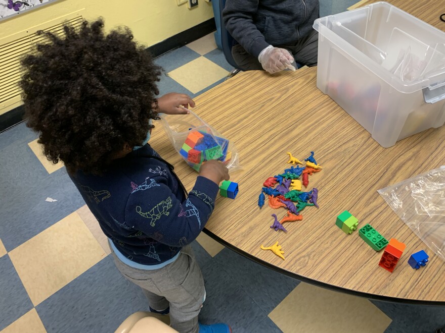 A 3-year-old boy puts blue and green blocks into a plastic bag. There are also purple, yellow, orange, green, red and blue dinosaurs on the table.
