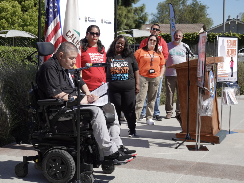 A man in a motorized wheelchair speaks into a microphone. People standing in the background listen.
