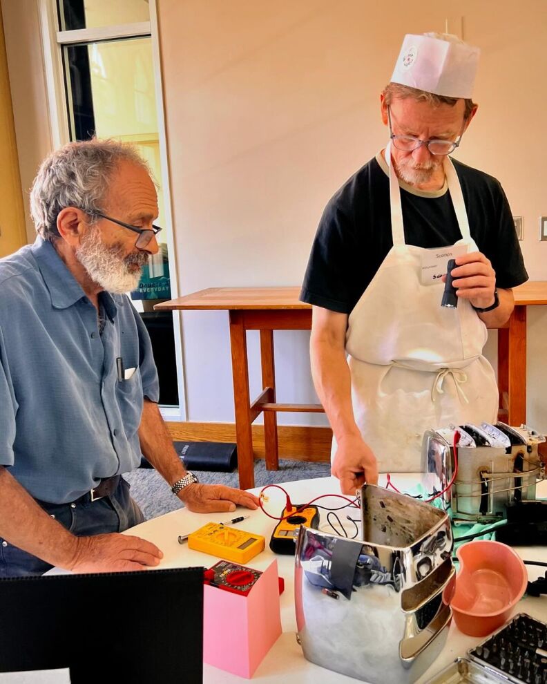 Two men stand at a table full of random gadgets and odd and ends. One is wearing a white apron and hat. 