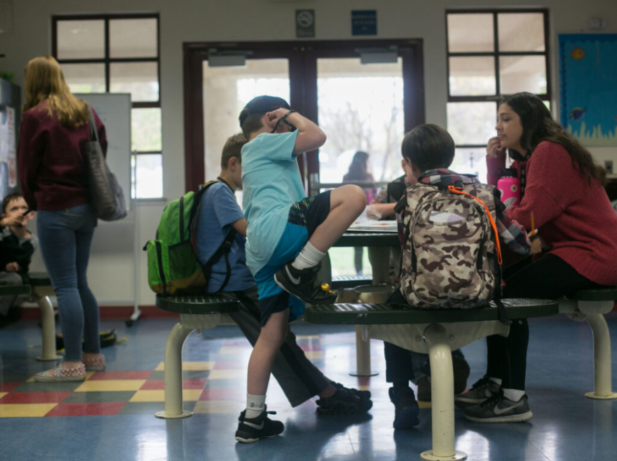 Students around 9 years of age and one older person are sitting at a table in a room next to doors with glass and windows near it.