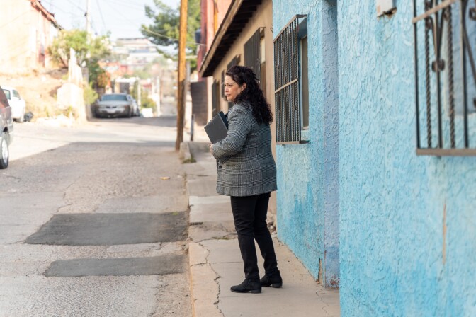 A middle-aged woman holding a binder stands on the street in front of a light blue wall. 