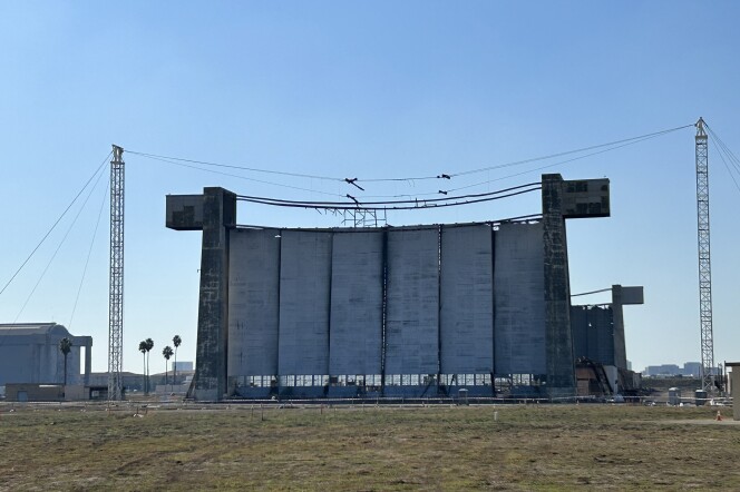  A view across a dirt and grass field at a wide, tall concrete-looking wall with six panels and concrete towers on either side. 