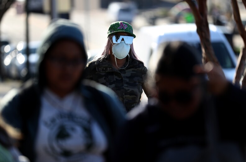 A person with a hat, sunglasses, and a mask on stands behind people at a distance. There are people in the foreground.