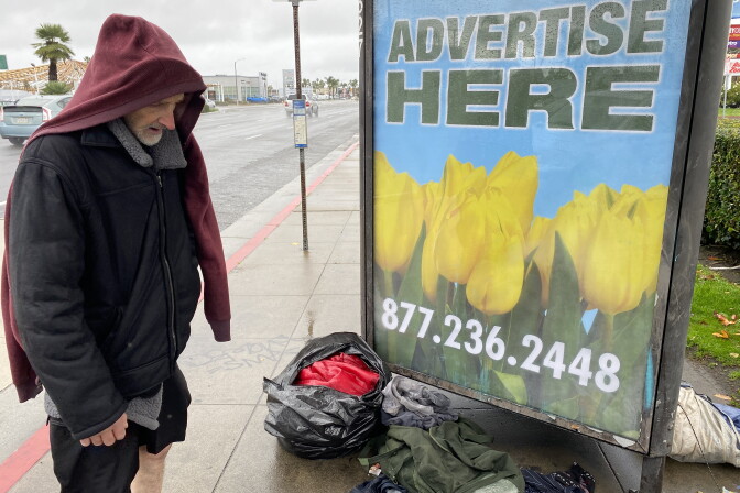 A man in a black jacket with a red hoodie over his head looks down at piles of clothing and a soaked red sleeping bag on the ground next to a road. A bus station advertising board is in the background. 