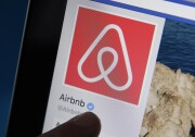 A close-up of a smart phone screen, specifically an Apple iPhone. A red and white company logo takes up most of the screen, with the text "Airbnb" below it in black with a small light-blue inverted checkmark next to it. The tip of a person's finger can be seen below the checkmark.