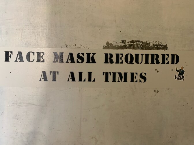 A sign stenciled in black on a parking garage wall reads, "Face mask required at all times." Just below the message is a small sticker that is a photograph of President Biden pointing to the sign’s message above the words, “I did that!” 