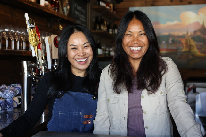 Two women both with medium dark skin stand behind a bar, smiling broadly at the camera. The woman on the left has long dark hair and wears a black top under navy denim overalls with leather strap details. The woman on the right has long dark hair and wears a light gray jacket over a purple top. Beer taps, wine glasses, and a mural depicting a Thai temple scene are visible in the background.