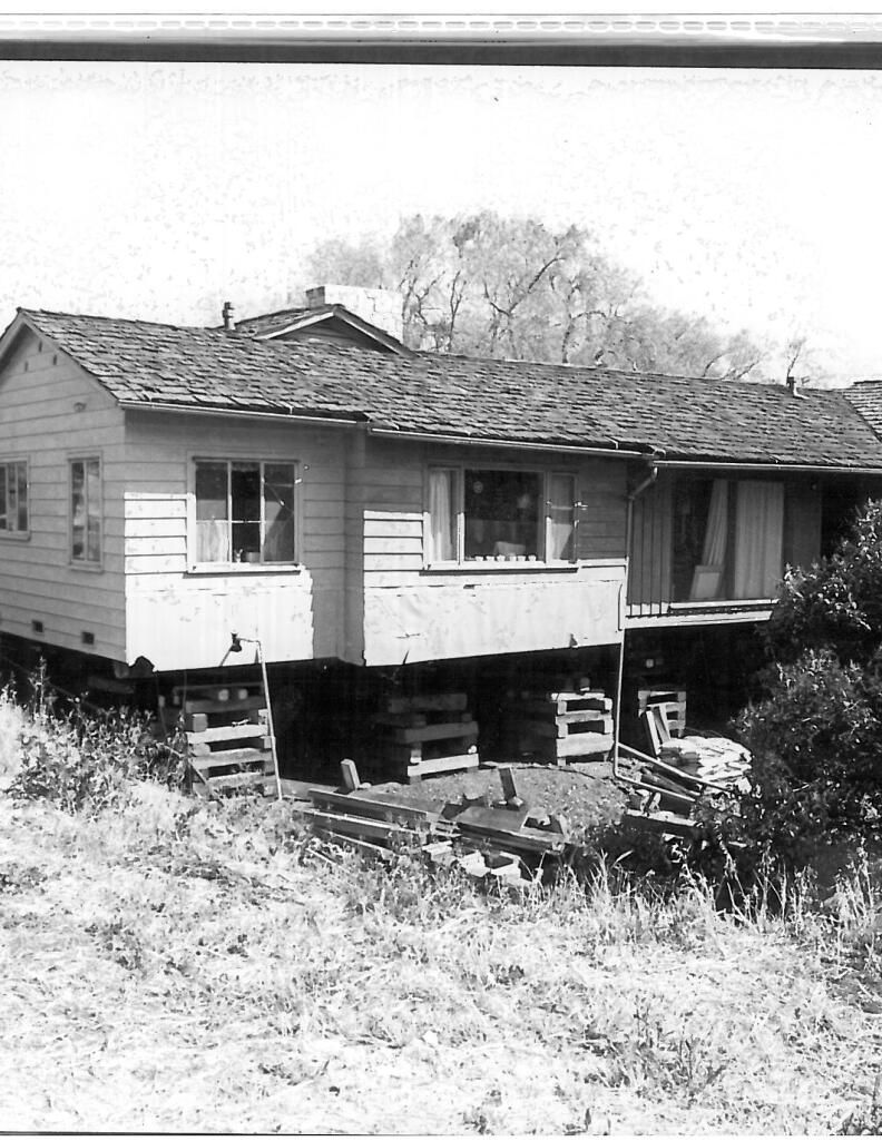 A black and white photo of a white single story house on stilts.